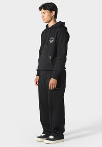 Czarna bluza z kapturem, z przednią kieszenią i sznurkiem, z złotą haftowaną aplikacją. W zestawie z czarnymi spodniami dresowymi z akcentem w postaci logo. Kanwasowe trampki.