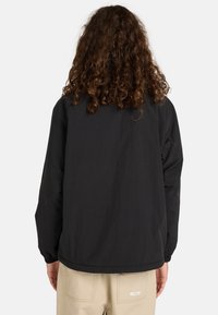 Zwarte lichte jas met een ontspannen pasvorm. Functies elastische manchetten en een ribbelzoom. Materiaal lijkt glad met minimale details.