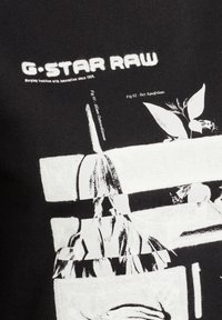 T-shirt en coton noir avec imprimé graphique blanc comprenant des motifs botaniques et du texte. Le design inclut des formes et des lignes abstraites, créant un contraste.