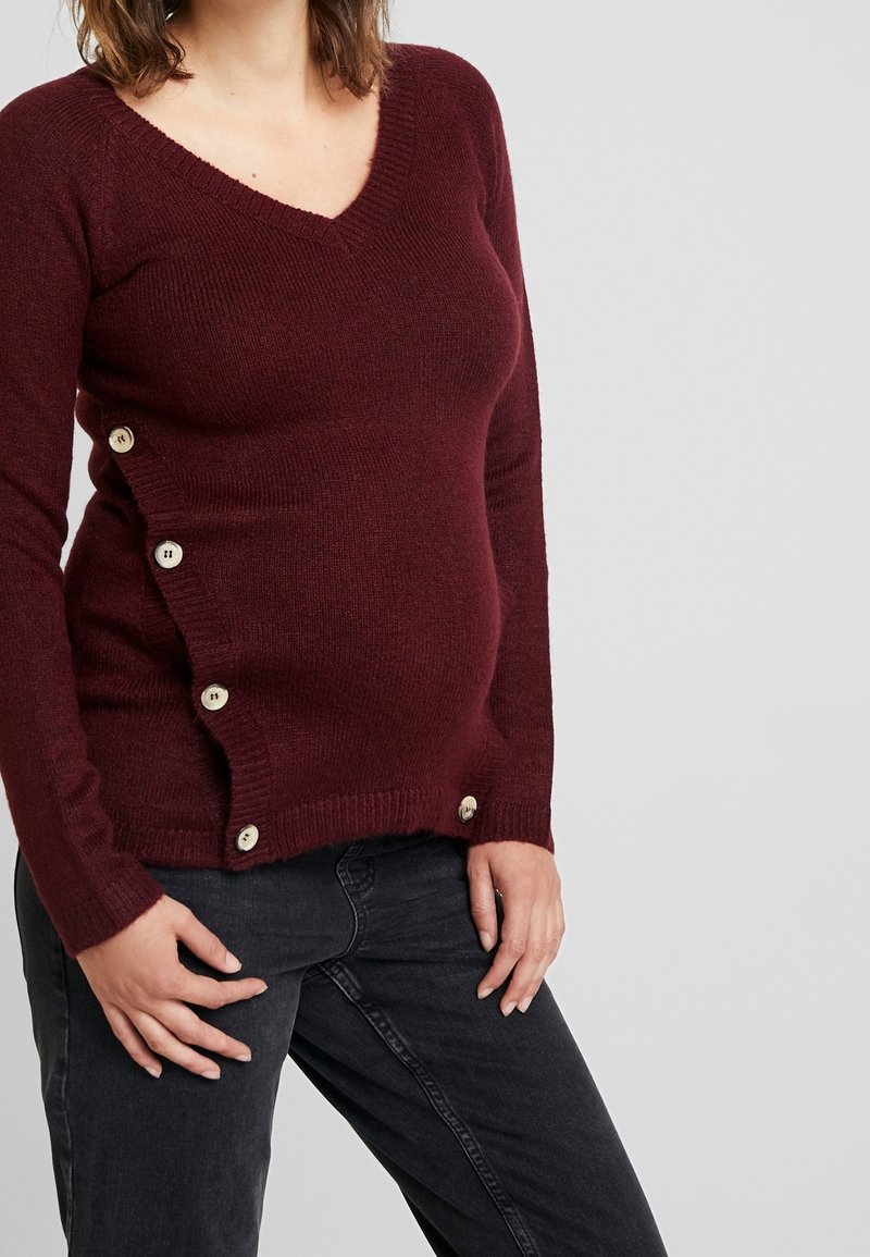 Femme enceinte portant un pull en maille bordeaux avec des boutons sur le côté et un jean noir, debout devant un fond clair uni.