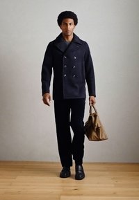 Dunkelblauer, doppelt geknöpfter Mantel mit großen Knöpfen, schwarze Hose und schwarze Schuhe; hält eine braune Tasche und steht auf einem Holzboden.