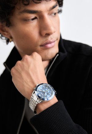 Reloj de metal plateado con esfera azul y correa de acero inoxidable, que cuenta con tres subesferas y un bisel giratorio con marcas.