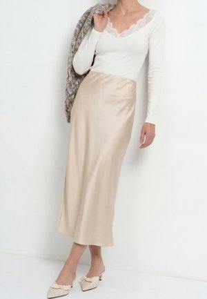 Frau trägt ein weißes langärmeliges Top mit Spitzenbesatz, beigen Satinrock, beige spitze Absatzschuhe, hält einen grob gestrickten Pullover über der Schulter.