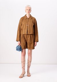Bruine suède cropped jack met zilveren knopen, bijpassende shorts en beige sandaletten met bandjes. Houdt een kleine blauwe handtas vast. Minimalistisch ontwerp.