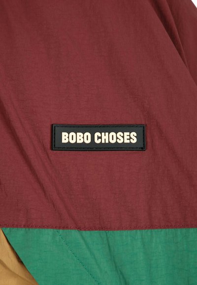 Étiquette rectangulaire noire avec texte blanc "BOBO CHOSES" sur tissu grenat au-dessus de sections de tissu vertes et beiges.