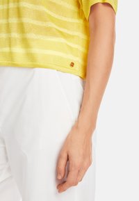 Gelb gestreifter Pullover aus leichtem Strickstoff, mit kurzen Ärmeln und einem goldenen Akzent am Saum. Kombiniert mit weißen Hosen.