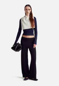 Zkrácená bílá mikina přes tmavě modrý dlouhý rukáv, doplněná černými volnými kalhotami. Modelka drží černou tašku s hladkou texturou.