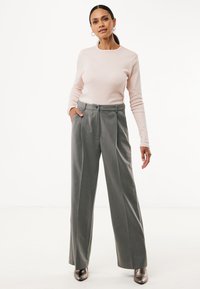 Frau mit dunklem Haar im Pferdeschwanz, die ein hellrosanes Langarmoberteil, hochtaillierte graue weite Hosen und metallische Stiefeletten trägt.