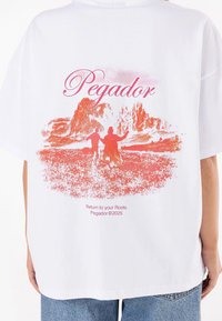 White cotton t-shirt featuring a large pink graphic print of mountains with figures, and text reading "Return to your Roots. Pegador ©2025."