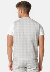Chaleco gris con un patrón y una textura suave, con corte recto y bolsillos traseros, llevado sobre una camiseta blanca.