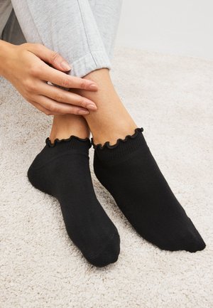 Lábak fekete boka zokniban fodros szegéllyel, világos szőnyegen, kezek óvatosan érintik az egyik bokát, személy világos szürke nadrágban.