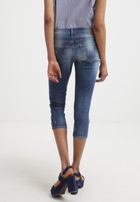 Dark blue denim capri jeans with a fitted design and frayed hem. Features two back pockets and light fading. Worn with chunky navy heels.