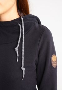 Kvinna som bär en svart hoodie med grå dragkedjor och en brun cirkulär patch på den övre vänstra ärmen, mot en vit bakgrund.