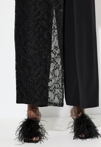 Vestido largo de encaje negro con un panel transparente, combinado con sandalias de piel de faux de color negro que tienen acentos de pom-pom texturizados, mostrando un diseño audaz.