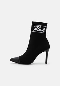 Bota estilo calcetín negra con un alto tacón de aguja, que presenta bordado de logotipo blanco y un diseño de punta afilada. Material superior de textil suave.