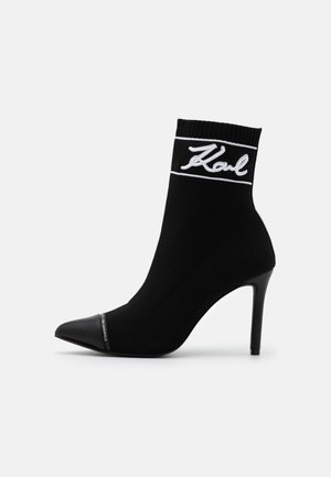 Czarne botki w stylu skarpet z wysokim obcasem typu stiletto, z białym haftem logo i spiczastym noskiem. Miękka tekstylna cholewka.