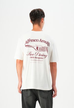 White cotton t-shirt featuring burgundy text and a fork graphic on the back. Short sleeves and relaxed fit; paired with dark gray jeans.