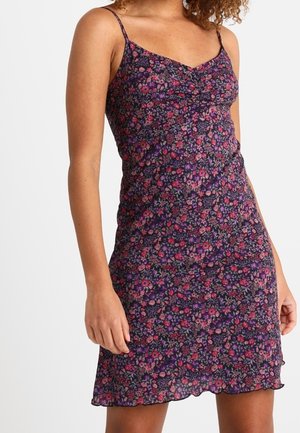 Mujer que lleva un vestido sin mangas, hasta la rodilla, con tirantes finos y un diseño floral oscuro con flores rosas y moradas.