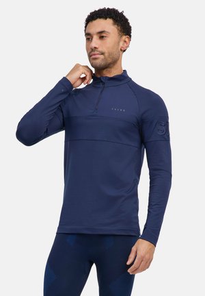 Mann trägt ein marineblaues, langärmliges Sportshirt mit Frontreißverschluss und passende Leggings, zieht mit der rechten Hand am Kragen.