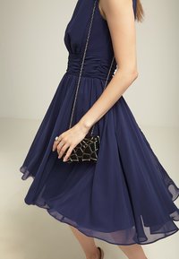 Granatowa sukienka bez rękawów z marszczoną talią, zwiewnym dół i przezroczystym materiałem. Para rąk trzyma czarną torebkę o strukturze z łańcuszkowym paskiem.