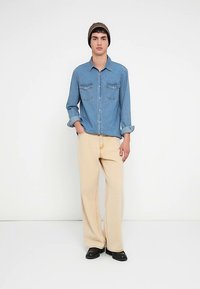 Blaues Denimhemd mit Knopfleiste und Brusttaschen, kombiniert mit beigen Hosen mit weitem Bein und schwarzem Schuhwerk. Eine Mütze sorgt für zusätzliche Details.