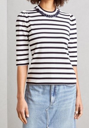 Vrouw draagt een zwart-wit horizontaal gestreept shirt met driekwartmouwen, een dikke zwarte ketting en een lichtblauwe denim rok.