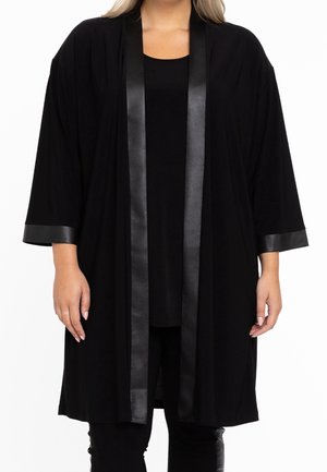 Femme portant un long cardigan noir ouvert sur le devant avec une bordure brillante le long des bords et des poignets des manches, associé à un pantalon noir.