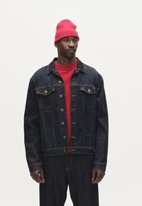Jeans jakna v temnem pranju s bronastimi gumbi, dvema žepoma na prsih, strukturirano prileganje; prekrita z svetlo rdečo pleteno majico in usklajeno kapo.