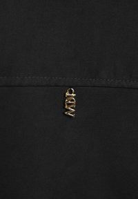 Zwarte stof met zichtbare stiksels en een klein gouden metalen label met de letters "MW" dat onder de naad is bevestigd.