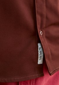 Καφέ βαμβακερό πουκάμισο με υφή, διαθέτοντας κουμπί κλεισίματος, αντίθετη ραφή και εμφανή ετικέτα Carhartt στη ραφή.