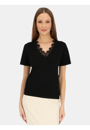 Kobieta ubrana w czarną koszulkę z krótkim rękawem z koronkowym dekoltem w kształcie litery V oraz jasnobeżową spódnicę, stojąca na jednolitym tle.