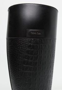 Bolsa de cubo de cuero negro con un patrón de cocodrilo texturizado, borde liso y un parche de logo negro. Acentos plateados completan el diseño elegante.