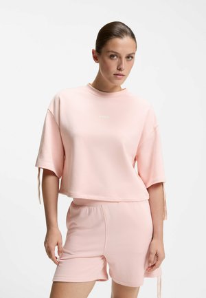 Vrouw draagt een lichtroze oversized top met korte mouwen en bijpassende short, staand tegen een effen witte achtergrond.