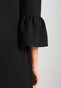 Svart klänning med en strukturerad tyg. Ärmen har en klockformad design med en volangkant, vilket visar en smidig övergång till en figurnära siluett.