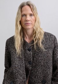 Chaqueta de lana texturizada con patrón de espiga marrón, que presenta un diseño de doble botonadura con grandes botones negros y un cuello con muesca.