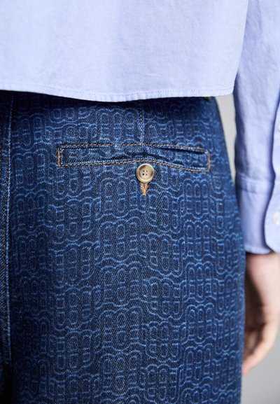 Κοντινό πλάνο της πίσω τσέπης σε μπλε μοτίβο τζιν με κούμπωμα, φορεμένο με ανοιχτό μπλε πουκάμισο.