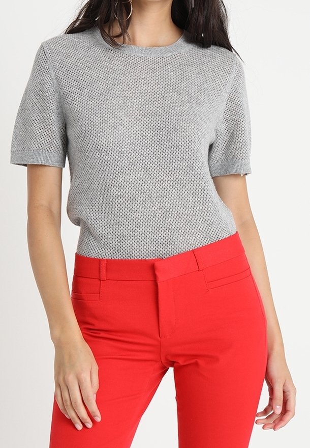 Femme portant un haut en maille gris clair à manches courtes et un pantalon rouge vif ajusté, debout, une main reposant le long du corps.