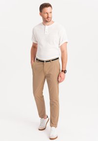 Top corto bianco testurizzato a maniche corte con tre bottoni, pantaloni chino beige e scarpe da ginnastica bianche con dettagli marroni. Outfit casual, design aderente.