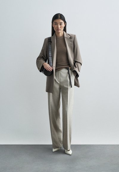 Καφέ κομψό σακάκι, μπεζ εφαρμοστό τοπ, ανοιχτό γκρι παντελόνι με φαρδιά πόδια και μαύρη ζώνη, και λευκά μυτερά παπούτσια. Μεσαίου μήκους σκούρα μαλλιά.