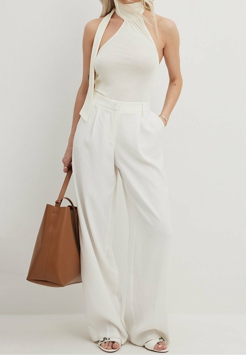 Mujer vestida con una blusa sin mangas blanca y pantalones de pierna ancha, sosteniendo un bolso tote marrón con una mano en el bolsillo.