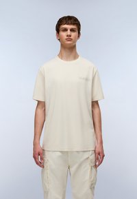 T-shirt en coton crème avec un col rond, des manches courtes et un logo subtil sur la poitrine gauche. Associé à un pantalon cargo léger.