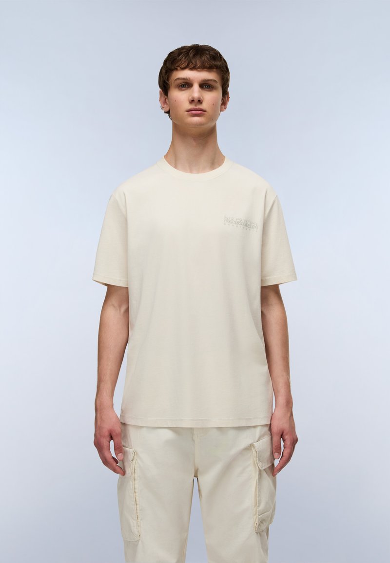 T-shirt en coton crème avec un col rond, des manches courtes et un logo subtil sur la poitrine gauche. Associé à un pantalon cargo léger.