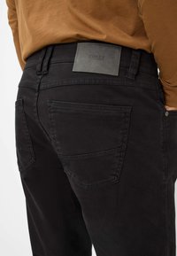 Zwarte katoenbroek met een gestructureerd leren logopatch op de tailleband, voorzien van twee achterzakken en zichtbare stikseldetails.