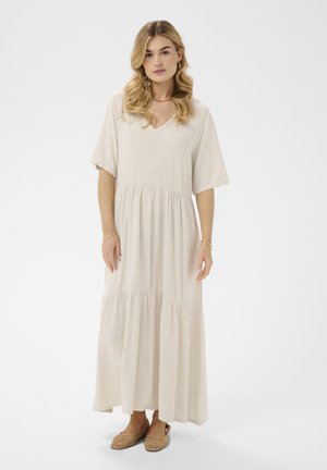 Szőke hajú nő, laza, világos bézs maxi ruhában és fonott barna bebújós cipőben áll tiszta fehér háttér előtt.