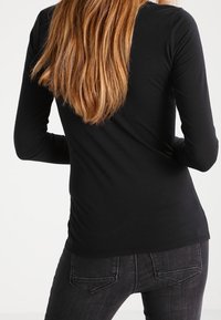 Schwarzes Langarmoberteil mit figurbetontem Design und glatter Textur, kombiniert mit dunklen Jeans. Der Rücken zeigt einen schlichten Ausschnitt ohne Verzierungen.