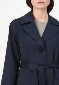 Navy blauwe trenchcoat met grote revers, knoopsluitingen en een strikriem in de taille. Gladde stof met een subtiele textuur.