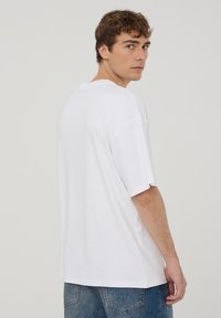 T-shirt bianca oversize in cotone, con maniche corte e scollo rotondo, abbinata a jeans in denim blu, vista da dietro.