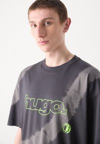 Tamsiai pilka oversize marškinėliai su džinsiniu raštu, dideliu žaliu "hugo." logotipu priekyje ir mažu apvaliu logotipo akcentu apačioje.