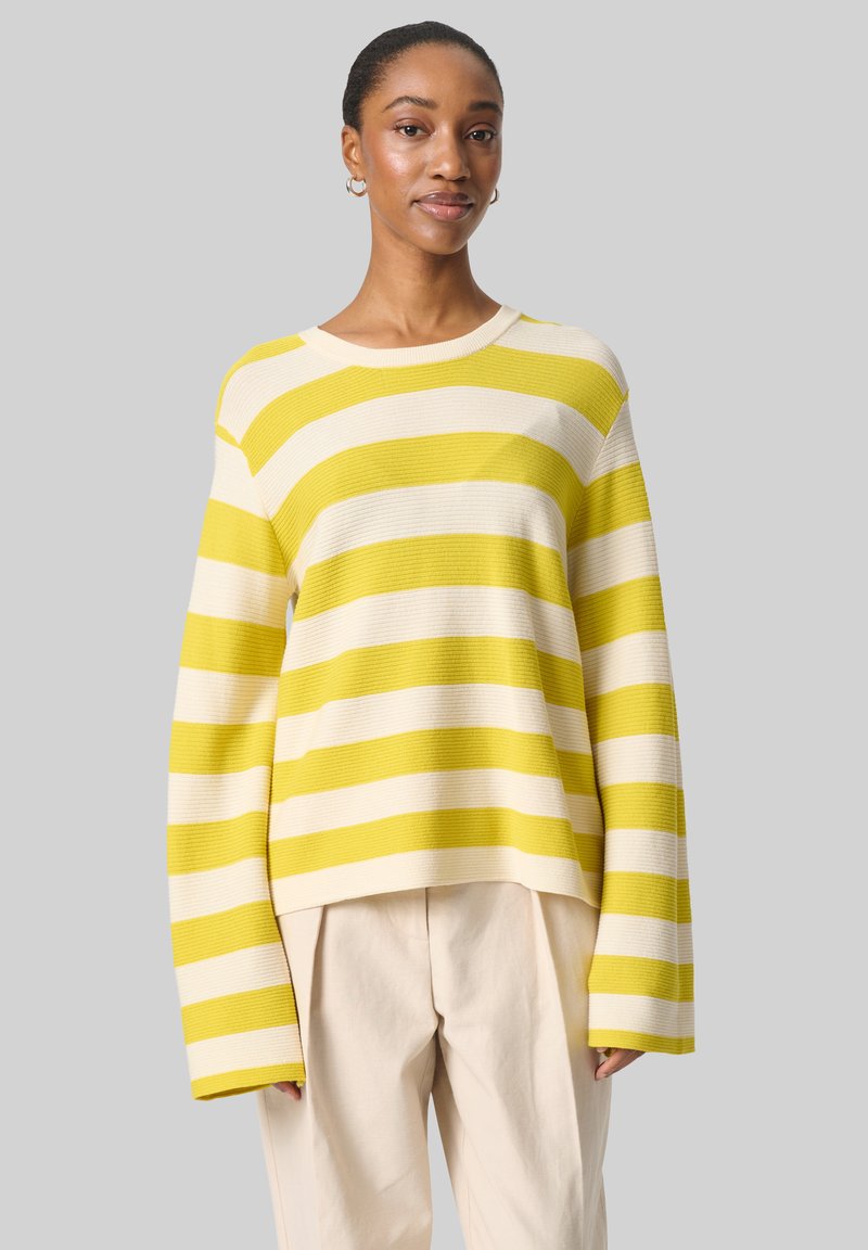 Pull rayé jaune et crème avec une texture côtelée, des manches larges et une coupe décontractée. Associé à un pantalon léger et ample.
