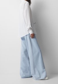 Camicia bianca a maniche lunghe con vestibilità ampia e orlo curvo, abbinata a pantaloni in denim a gamba larga di un blu chiaro, mostrata in vista laterale.